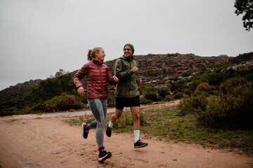 Young fit couple laughing while running on gravel mountain track in cloudy weather with luscious bushes