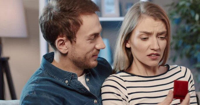 Young couple sitting at home on couch. Man gives woman box with wedding ring, dissatisfied woman refuses him.