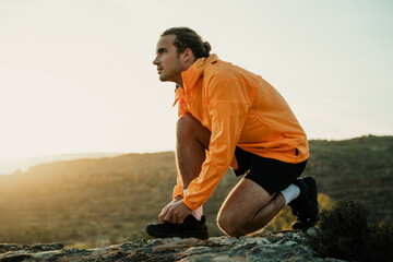 Caucasian male athlete taking a break from run tying shoe lace while hiking in mountain during sunset