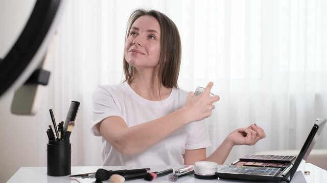 Woman blogger doing make-up at home, apply perfumes, smelling aroma. Beautiful young female near the mirror in the living room, self care and product presentation for users online 