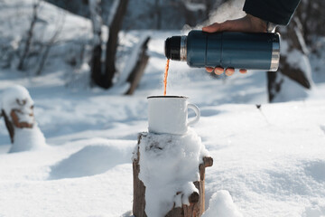 Male hand pouring hot tea into a mug from a thermos in the winter forest. Warming drink in cold weather. Hike, camping concept.