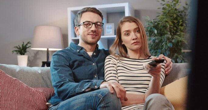 Young couple sitting at home on couch and watching movie. Two people spend free time together. Woman constantly switches channels using the remote control.