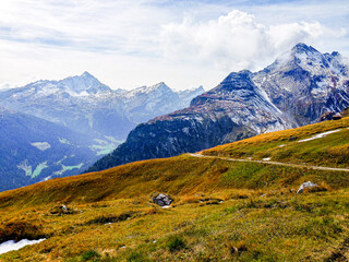 landscape in the mountains with snow on mountain peaks and green field and a small road going there in winter or autumn