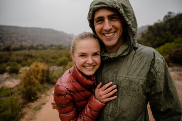 Happy smiling couple resting after steep trail run up gravel mountain slope in cold cloudy weather with rain jackets