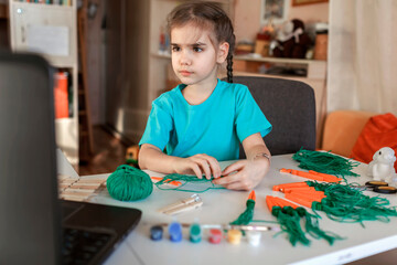 Elementary school girl doing carrot with wooden clothes pins for Easter celebration looking video on laptop, online workshop, kid at-home activity, creativity and distant education, holiday decoration