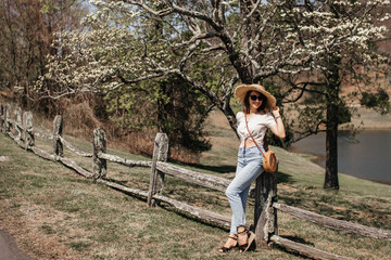 A beautiful girl with brown curly hair, sunglasses and a straw hat, linen cropped blouse with a knot and jeans, stands near an old fence under flowering dogwood trees on a sunny spring day.