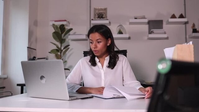 Businesswoman having online talk and interviewing at table with laptop in company spbi. Young woman looking at computer screen and talking, taking notes on paper resume and sitting at desk in light