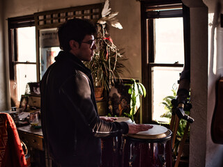Side view of young guy in stylish jacket and glasses playing bongo drums and looking away in cozy...