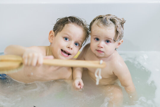 Funny little girl and her cute brother having fun taking a bath together playing in water with foam with colorful toys after a shower.