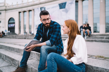 Fototapeta na wymiar Caucasian male and female digital nomad resting at city stairs discussing freelance project during travel vacations, youthful tourists with laptop talking during leisure time for communicate