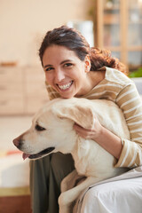 Vertical warm toned portrait of smiling young woman hugging dog sitting on bed in cozy home interior
