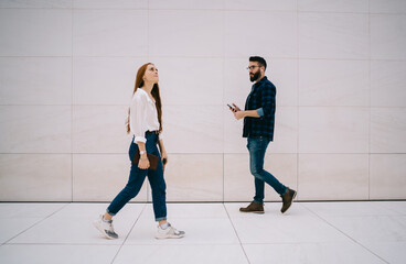 Side view of red hair female student with textbook walking in parallel of Caucasian millennial guy in electronic earphones listening music playlist from media app, publicity area at urban setting