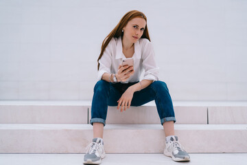 Portrait of attractive Caucasian woman with modern cellphone device resting at publicity area and looking at camera,millennial hipster girl with smartphone posing at stairs with promotional background