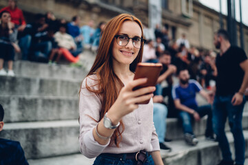 Half length portrait of cheerful female blogger in eyeglasses holding cellphone gadget smiling at camera enjoying solo travelling, happy Caucasian woman with red hair using smartphone technology