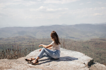 Naklejka na ściany i meble A beautiful girl with brown curly hair, in a linen short blouse with a knot and jeans, sits on a bald stone and looks into the mountains on a sunny spring day.