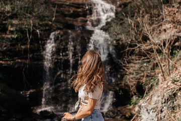 A young girl with brown curly hair in blue jeans and a linen sweater looks at a waterfall on a spring sunny day