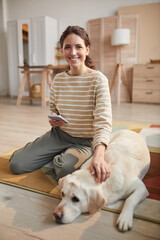 Vertical full length portrait of smiling young woman looking at camera while sitting on floor with pet dog in cozy home interior