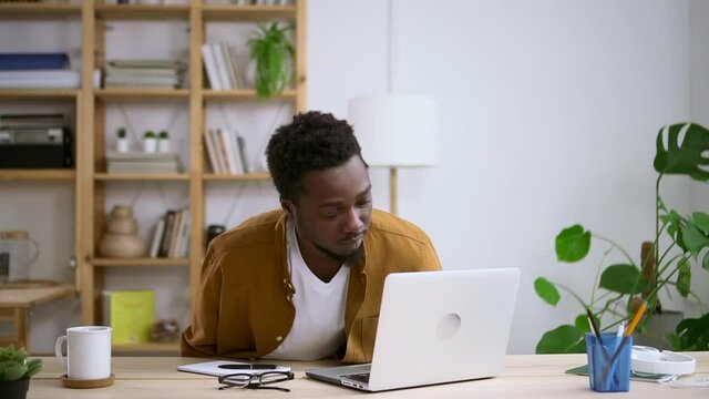 Man working on startup project and sitting at desk at home room during pandemic spbas. Young businessman sits down at table, looks at computer screen and types in light room. American African guy does