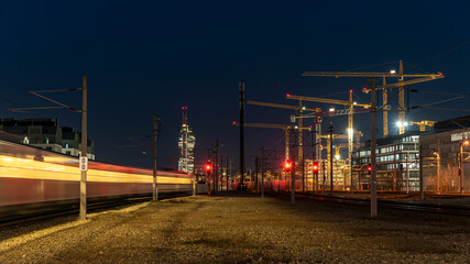 Fototapeta na wymiar Langzeitbelichtung Zug fährt in der Nacht in die Station ein. Im Hintergrund Kräne einer Baustelle und ein Wolkenkratzer. Zugsignal zeigt rot.