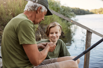 Man fisherman teaching his grandson to hook bait, renowned as method for catching fish, smiling young blonde guy looks at senior male with smile and concentrated look, sit on wooden stairs to water.