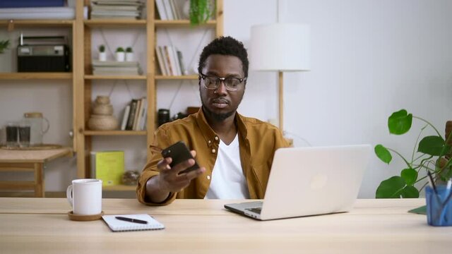 Man using laptop and smartphone and sitting at table in home office spbas. Young businessman looks at computer screen then takes phone in hands and types, sits at desk in light room. African American