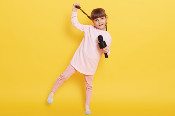 Fototapeta na wymiar Little girl with microphone standing on one leg isolated over yellow background, pushing one pigtail aside, dancing and performing concert, attractive little vocalist.