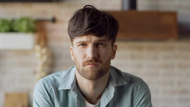 Head and shoulders portrait of sad young man with beard raising eyes and looking at camera with blank facial expression at home