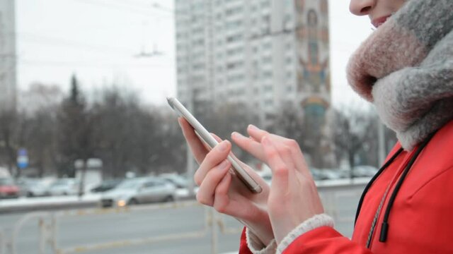 Close up of woman hands texting on smartphone standing on street in winter city against the background of a road with moving cars