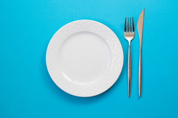 Empty white plate, fork and knife on blue background. Top view.