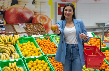 A smiling woman is buying fruits in the supermarket and showing an apple to the camera. 