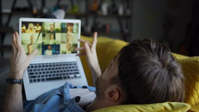 Tracking over the shoulder shot of young man having video call on laptop with group of defocused colleagues or business partners lying on sofa at home