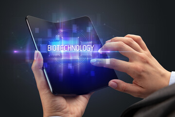 Obraz premium Businessman holding a foldable smartphone with BIOTECHNOLOGY inscription, new technology concept