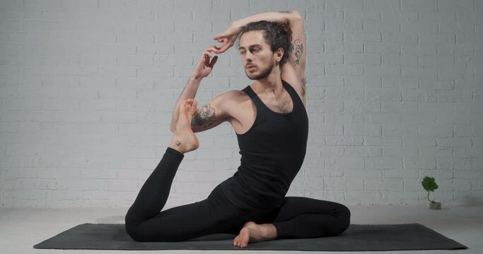 Young sporty man practicing yoga, doing Mermaid exercise, Eka Pada Rajakapotasana pose, working out, wearing sportswear, pants and top, indoor, white yoga studio
