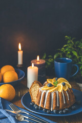 Orange bundt cake with blueberry surrounded fruits, candles, plant and cutleries on dark wooden background.