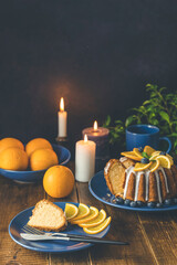 Orange bundt cake with blueberry surrounded fruits, candles, plant and cutleries on dark wooden background.