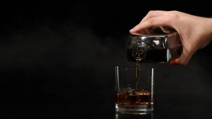 Barman pour golden whiskey, cognac, brandy from bottle into drinking glass with ice cubes on black dark background. Bartender pouring tasty alcohol cocktail drink bourbon, rum in slow motion. Close-up