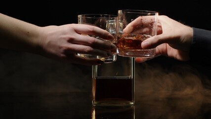 Two hands with glasses of cognac whiskey with ice cubes making cheers on black background....