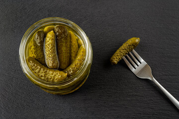 Open glass jar of tasty pickled gherkins and one more on the spoon against black slate background....