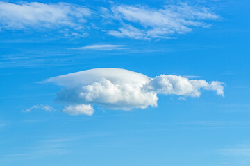 Small lenticular cloud against translucent cirrus clouds high in a clear blue sky. Different cloud types and atmospheric phenomena. Skyscape on a sunny day. Meteorology and weather.