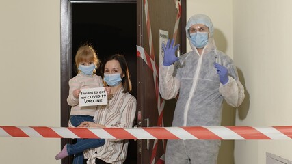 Medical worker in suit with text on paper visiting young family of mother and daughter at home during coronavirus Covid-19 quarantine lockdown. Doctor opening door with warning tapes in entrance hall