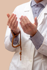 A young man in a white robe raised his hands up and prayed with a Muslim rosary. Beige background. Doctor praying. A Muslim makes dua.