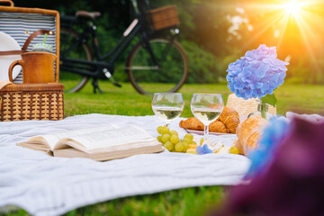 Summer picnic on sunny day with bread, fruit, bouquet hydrangea flowers, glasses wine, straw hat, book and ukulele. Picnic basket on grass with food and drink on white knit blanket. Selective focus