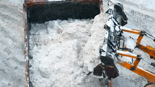 Close-up overhead above view of tractor loader machine snowplow shovel loading pile of snow and ice in dump truck for removal from city street at blizzard snowfall. Snow cleanong and removal