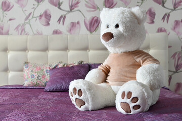 Big white teddy bear toy is sitting on the bed in purple bedroom interior.