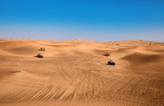 Beautiful Dubai desert landscape with plants and two riding quad buggy vehicles, sand with wheel tracks and blue sky