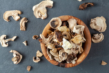 Close up isolated flat lay image of a rustic wooden bowl filled with sliced and dried mushroom pieces. The bowl is on dark stone surface. There is an assortment of mushroom types in this moody image.