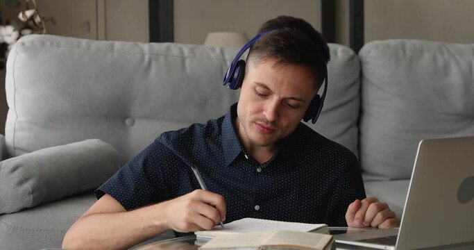 Focused young handsome man in headphones listening educational lecture or online webinar in computer application, writing notes in copybook, improving knowledge distantly at home. e-learning concept.
