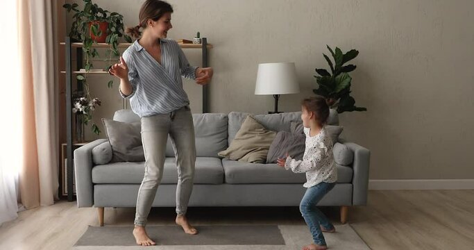 Full length joyful young mother dancing to disco music barefoot with cute energetic little preschool kid daughter on floor carpet, involved in crazy weekend domestic activity together at home.