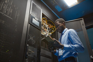 Low angle of African American man holding digital tablet while standing by server cabinet and...