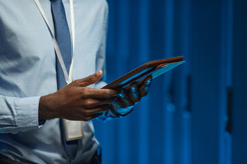 Cropped shot of African American data engineer holding digital tablet while working with supercomputer in server room lit by blue light, copy space